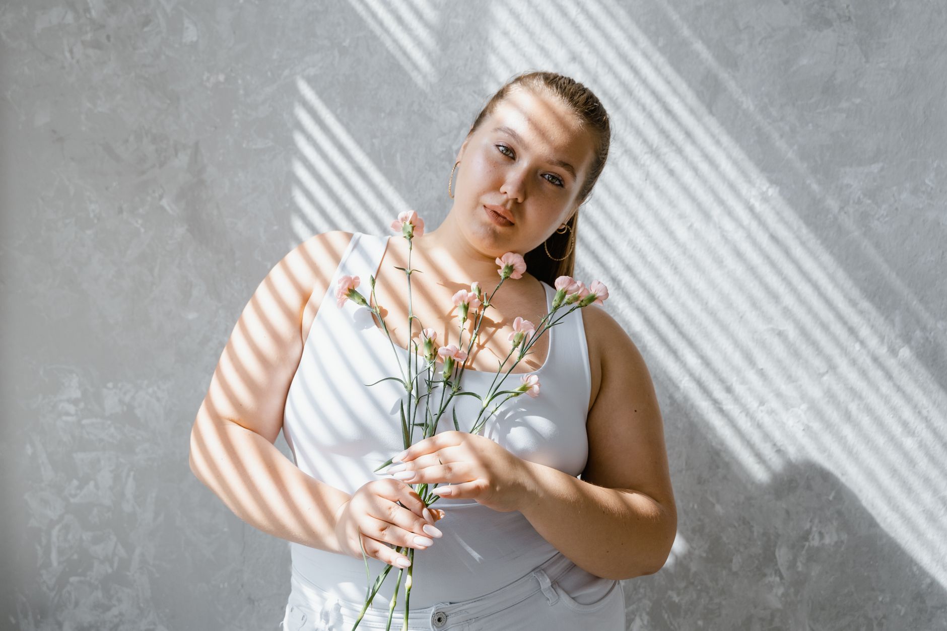 Uśmiechnięta kobieta plus size w topie trzymająca kwiaty w świetle żaluzji Uśmiechnięta kobieta plus size w topie trzymająca kwiaty w świetle żaluzji